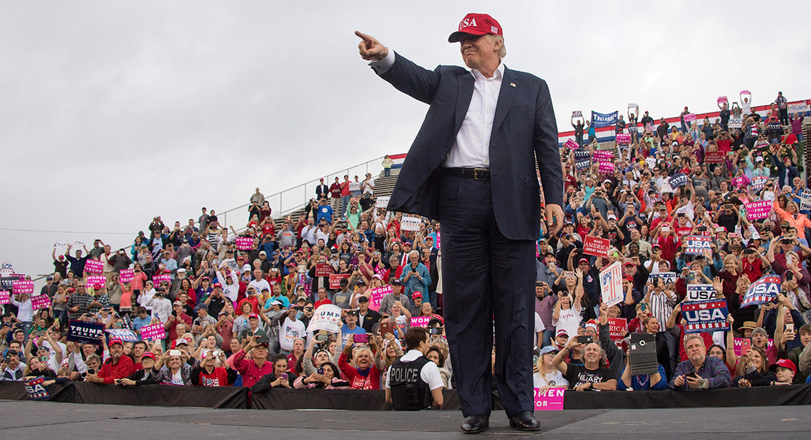 Donald Trump stands on a stage pointing toward a packed outdoor rally crowd holding campaign signs.